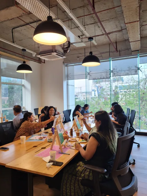 Colleagues seated around a boardroom table with mini easels and craft kits at their places, a Heart Arts India facilitator leading the session from the head of the table