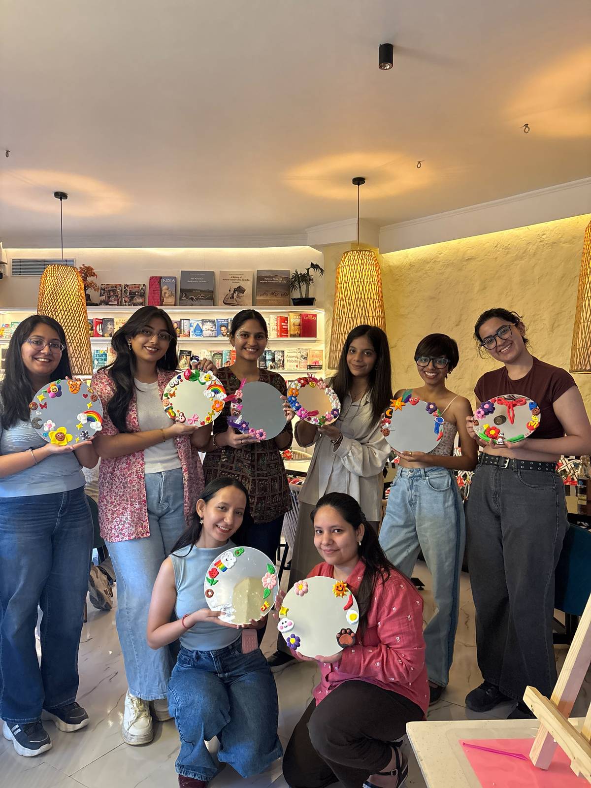 Eight participants at a Heart Arts India workshop holding up the plates they decorated — hand-painted flowers, rainbows and clay figures on each one.